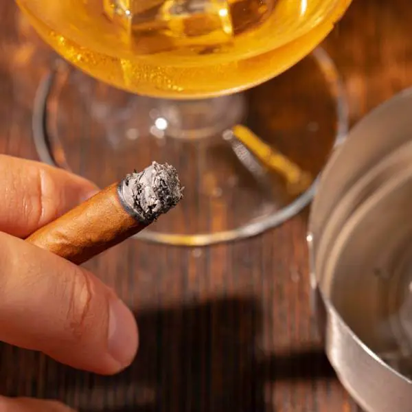 Une main tenant un cigare près d'un verre d'alcool.